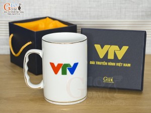 Cốc sứ dáng cao quai nắm men trắng kẻ chỉ vàng kim, in logo VTV 450ml